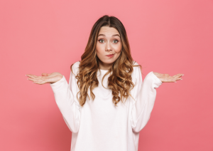 Female looking confused shrugging, wearing a white top against a pink wall