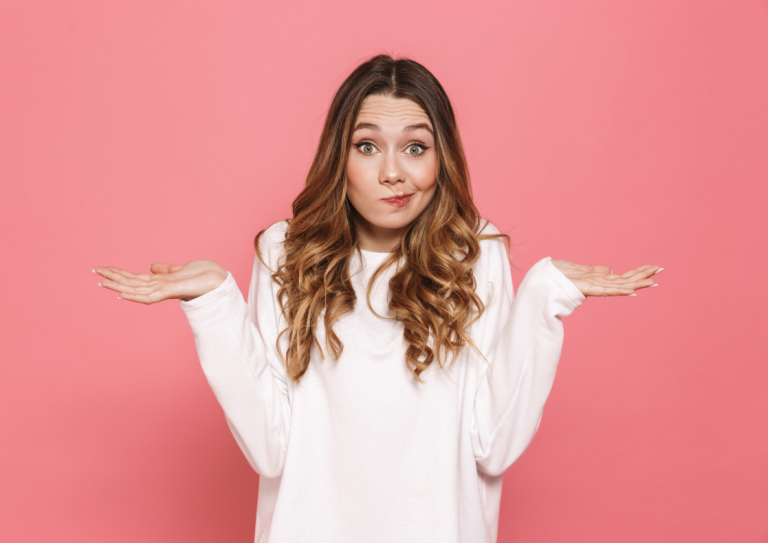 Female looking confused shrugging, wearing a white top against a pink wall